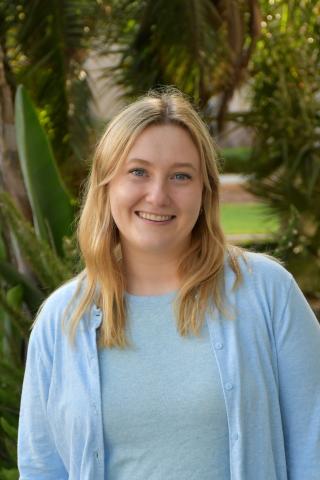 Laura smiling and wearing a baby blue shirt and cardigan, greenery and trees in the background